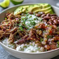 Golden brown, crispy carnitas pile high over fluffy white rice in a wide bowl, topped with creamy avocado slices and vibrant fresh salsa.